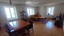 A spacious living and dining area with wooden flooring, large windows, a sofa, coffee table, dining table with chairs, and a shelf with decorative items.