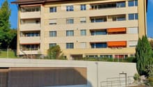 Four-story residential building with beige facade, multiple windows, balconies, and a fenced parking area at the base.
