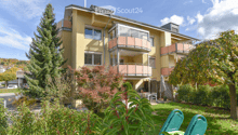 A three-story yellow building with multiple balconies and large windows, surrounded by greenery and a small patio area with outdoor furniture.