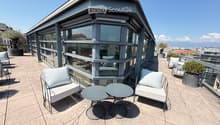 Modern rooftop terrace with a sofa, two round tables, and several chairs, surrounded by glass walls and a cityscape view.