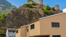 Modern two-story building with beige facade, large garage doors, and a well-maintained lawn, situated in front of a rocky hill with a castle-like structure on top.