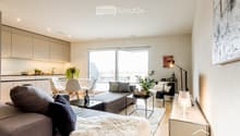 Modern open-plan living and dining area with a grey sofa, coffee table, armchair, ottoman, floor lamp, dining table with chairs, kitchen cabinet, faucet, plants, picture frame, bottle, and wine glass.