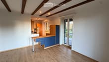 Bright, empty room with wooden flooring, white walls, and exposed wooden beams on the ceiling. Features a sliding glass door leading to an outdoor area and a small kitchen area visible in the background.