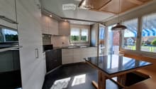 Modern kitchen with light-colored cabinets, a central island, and a dining area with a black countertop table and wooden chairs. The space features large windows and a pendant light hanging from the ceiling.