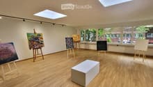 An art studio with wooden floors, large windows, and several easels displaying paintings. A white stool is placed in the center of the room.