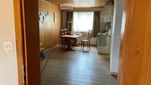 A furnished room with a wooden table and chairs, a kitchen area with a stove and fridge, and wooden walls and flooring.