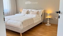Modern bedroom with a large bed, white bedding, wooden floor, nightstand with lamp, and sheer curtains.