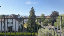 Balcony with wooden decking and metal railing, overlooking a residential area with houses, trees, and a clear blue sky.