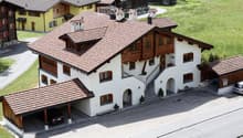 A large, two-story house with a tiled roof, white walls, and wooden accents. Features include a garage, a driveway, and surrounding greenery.