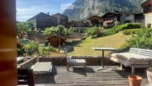 Wooden deck with outdoor furniture including a table, chairs, and a bench, overlooking a scenic mountain view with surrounding houses and greenery.
