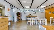 Bright, modern room with wooden flooring, white walls, and slanted ceiling. Features include a long wooden counter, several tables and chairs, a shelf with bottles, and a refrigerator.