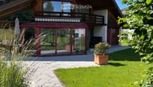 Modern two-story house with a sloped wooden roof, large windows, and a balcony. The house features a patio area with glass walls, a small garden, and a well-maintained lawn.