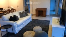 Modern living room with a white sofa, decorative pillows, a dark gray rug, three round woven stools, a small black side table, a white TV stand, and a dining area with chairs and a table visible in the background.