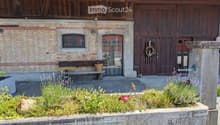 Garden area in front of a brick building with a large wooden door, featuring a bench, small table, flower pot, and decorative wreath on the door.