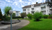 Well-maintained garden area with a circular stone fountain, neatly trimmed hedges, and a paved walkway leading to a multi-story residential building with balconies.