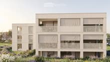 Modern two-story building with beige facade, large windows, and balconies. The ground floor has a raised entrance, and the building is surrounded by a well-maintained garden with various plants and flowers.