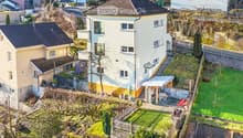 Aerial view of a two-story house with a sloped roof, surrounded by a garden and outdoor seating area. The house has multiple windows and a small staircase leading to the entrance. The garden includes a patio with a table and chairs, and there are addition
