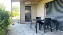 Outdoor patio with a wooden ceiling, tiled floor, and a black wicker dining set including a table and chairs. A sliding glass door leads to the interior, and there are potted plants and greenery nearby.