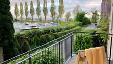 Balcony with wooden decking, metal railing, and a view of a harbor with boats and trees.