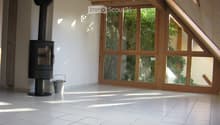 Bright, empty room with white tiled flooring, large wooden-framed windows, and a modern black wood-burning stove.