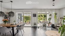 Modern dining area with a wooden table, grey chairs, pendant lights, shelves with bottles, a vase with flowers, and a cat tree. Large glass doors open to a patio with outdoor furniture and plants.