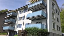 Modern multi-story residential building with white facade, blue shutters, and multiple balconies with glass railings and retractable awnings.