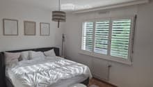 Bright bedroom with a large bed, white bedding, a small stool, radiator, and three picture frames on the wall. Large windows with blinds let in natural light.