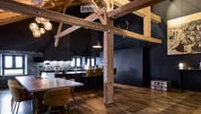 Open-concept dining and kitchen area with exposed wooden beams, wooden flooring, and modern lighting. Features a large wooden dining table with chairs, a kitchen island with stools, pendant lights, and a large picture frame on the wall.