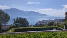 A scenic garden terrace overlooking a lake and distant mountains, with neatly trimmed hedges, outdoor chairs, and umbrellas.