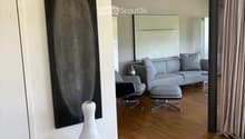 Modern living room with hardwood flooring, grey sofa, ottoman, chairs, a white vase on a table, and a large mirror on the wall.