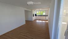 Empty room with wooden flooring and white walls, leading to a small kitchen area with white cabinets and black countertops.