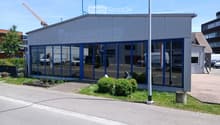Modern single-story building with large glass windows and blue framing, surrounded by a small landscaped area with shrubs and a paved walkway.