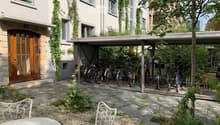 Garden area with stone-paved ground, metal chairs, a round table, bicycles under a covered area, and a building with white walls and large windows.