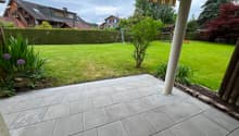 Well-maintained garden with a tiled patio, green lawn, hedge border, and surrounding trees and houses.