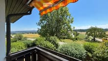 Balcony with wooden railing, striped awning, overlooking a rural landscape with fields, trees, and distant hills.