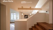 Open kitchen area with light-colored cabinets, wooden staircase, pendant lights, and large windows allowing natural light.