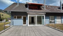 Two-story house with grey roof, light blue walls, large windows, and a covered balcony area with a railing.