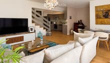 Modern living room with a white sofa, glass coffee table, and a TV stand. The room features a staircase leading to an open-plan kitchen area with modern appliances.