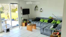 Bright living room with large sliding glass doors opening to a garden, grey sectional sofa with green cushions, wooden coffee table, cat tree, cat house, small table with flower pot, and a television mounted on the wall.