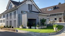 Modern two-story house with grey siding, multiple windows, and a garage entrance. The front yard features a well-maintained lawn, potted plants, and a small garden area.