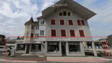 3 story building, gray exterior, red shutters, storefronts on the ground floor, sign 'wenger wohnen', sign 'Polsterarbeiten', sign 'Vorhänge Teppiche'