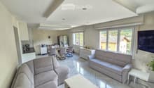 Bright living room with a grey sectional sofa, coffee table, dining area with chairs and table, kitchen area with refrigerator and cabinets, large windows with blinds, and a TV on the wall.