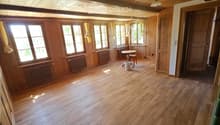Empty room with wooden flooring, wooden walls, large windows with yellow curtains, radiators, and a small round table with a chair near the windows.