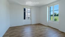 Empty room with white walls, wooden flooring, and large windows offering a view of a green field.