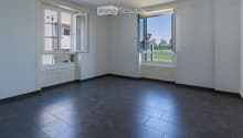 Empty room with dark tiled floor, white walls, and large windows with open shutters, showing a view of the outside area.