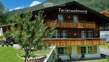 A two-story chalet with a balcony, surrounded by a well-maintained garden and a mountain backdrop.