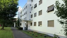 Modern multi-story building with white facade, multiple windows, and a small garden area with flowers and a tree.