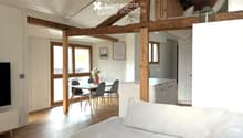 Bright, open-plan living and dining area with exposed wooden beams, large windows, a sofa, dining table with chairs, and a small kitchen area with cabinets and a refrigerator.
