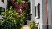 A narrow garden pathway lined with greenery leads to a white house with green shutters and a small balcony. The house has a textured facade and a visible address number30.