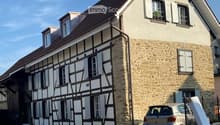 Two-story house with a combination of stone and timber framing, multiple windows with shutters, and a parked car in front.